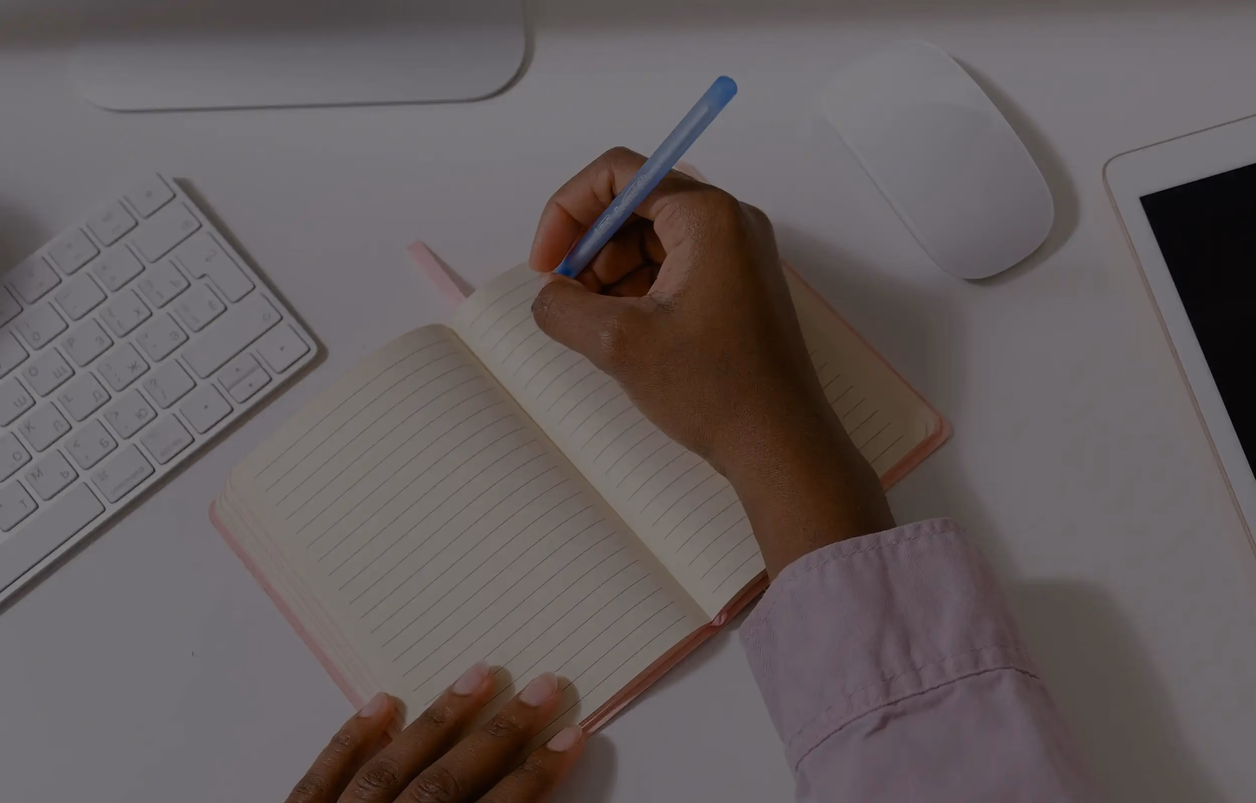 Eine Hand, die in ein kleines Notizbuch schreibt. Daneben liegt eine Mac Tastatur und ein iPad.
